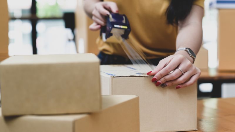 Someone seals a cardboard box with tape while other boxes are on the table around them.