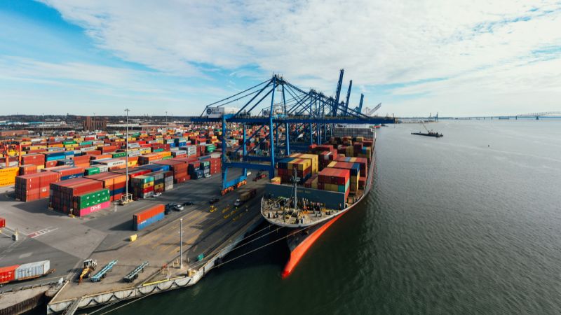 A busy port with colorful containers, large cranes, a docked cargo ship, and a partly cloudy sky.