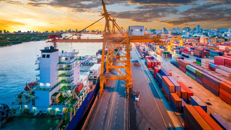 Cargo ship docked at port with large yellow cranes and shipping containers, bathed in golden sunset light.